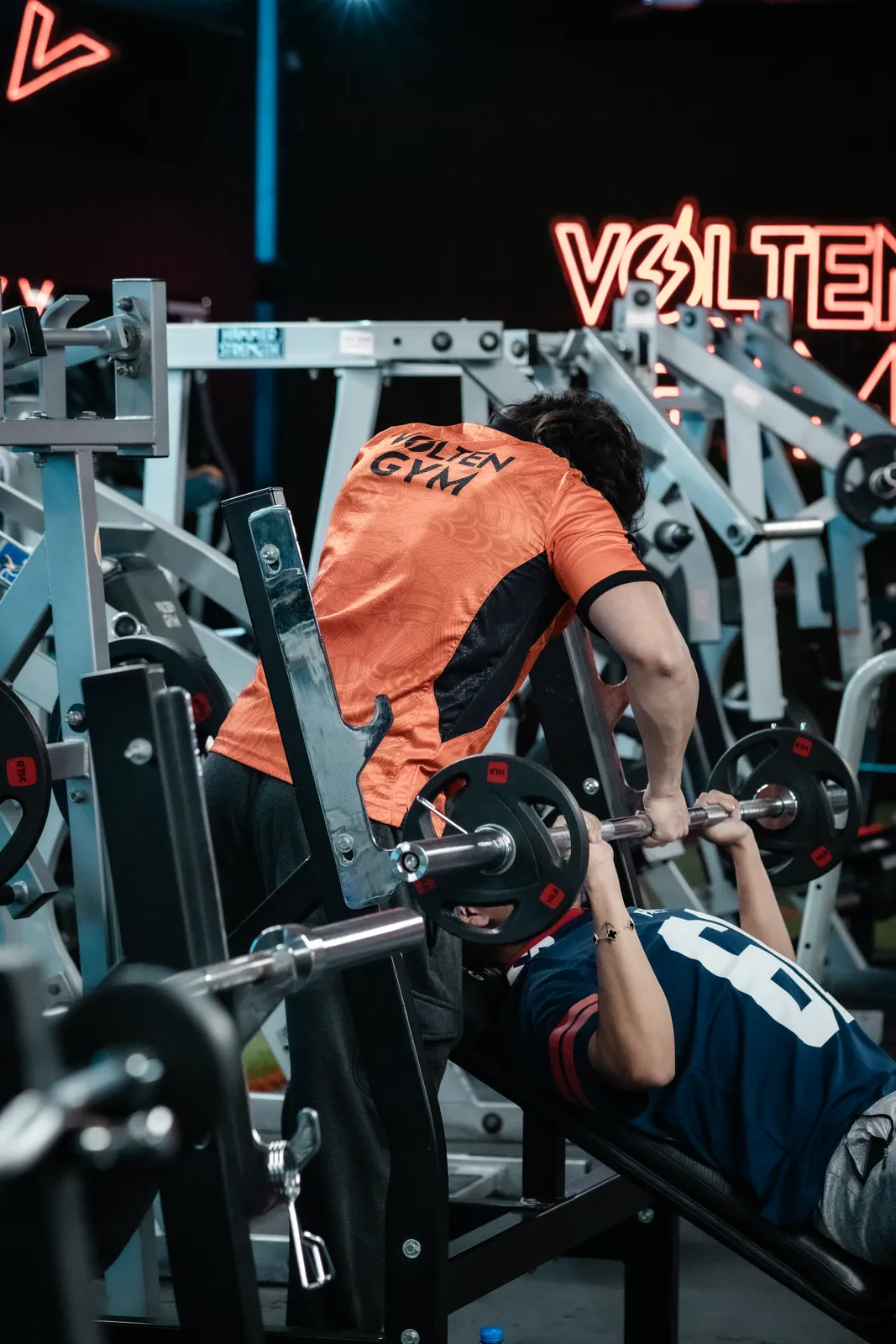 Socio con la playera Volten Gym haciendo press de banca, con el letrero de neón de Volten al fondo